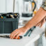 A handyperson in a plaid shirt and tool belt reaches for a pair of pliers on a white countertop. Nearby, an open toolbox holds various tools. Blurred in the background is a kitchen sink and a glass kettle. It's time to call in some expert help.